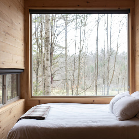 Gemütliches Schlafzimmer mit weißem Bett und Kopfkissen vor einem großen Panoramafenster mit Blick auf einen ruhigen Wald – einladendes Ambiente für erholsame Nächte und hochwertige Schlafkultur.