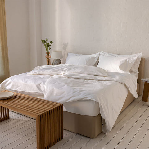 Ein einladendes Doppelbett mit weichem, weißem Excellence Bettdeckenbezug und Kissen in einem hellen Raum mit Holzdielen, einer rustikalen Holzbank am Fußende und einer Nachttischlampe.