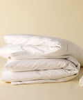 Ein fluffig zusammengelegter Stapel weißer Bettwäsche, bestehend aus Kissenbezügen und einem Excellence Bettdeckenbezug, neben einem verpackten Laken auf warmem, beigen Untergrund.