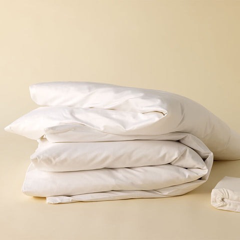Ein fluffig zusammengelegter Stapel weißer Bettwäsche, bestehend aus Kissenbezügen und einem Excellence Bettdeckenbezug, neben einem verpackten Laken auf warmem, beigen Untergrund.