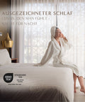 Frau sitzt entspannt auf dem Bett und genießt den Komfort des Excellence Gänsedaunenkopfkissens für erholsame Nächte und luxuriösen Schlaf.
